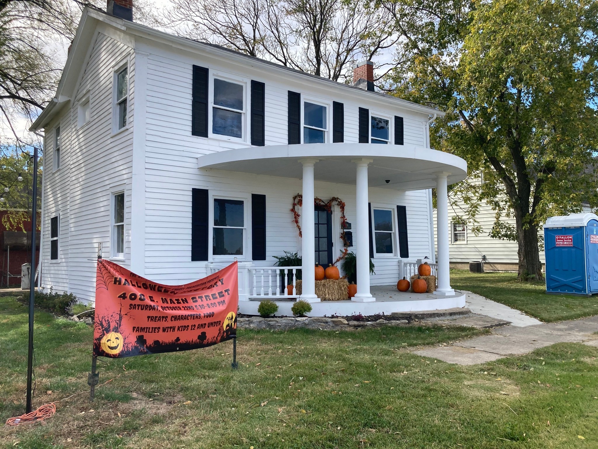 Two-story white house with black shutters, a curved porch with columns, pumpkins, and fall decorations, on a grassy lawn.
