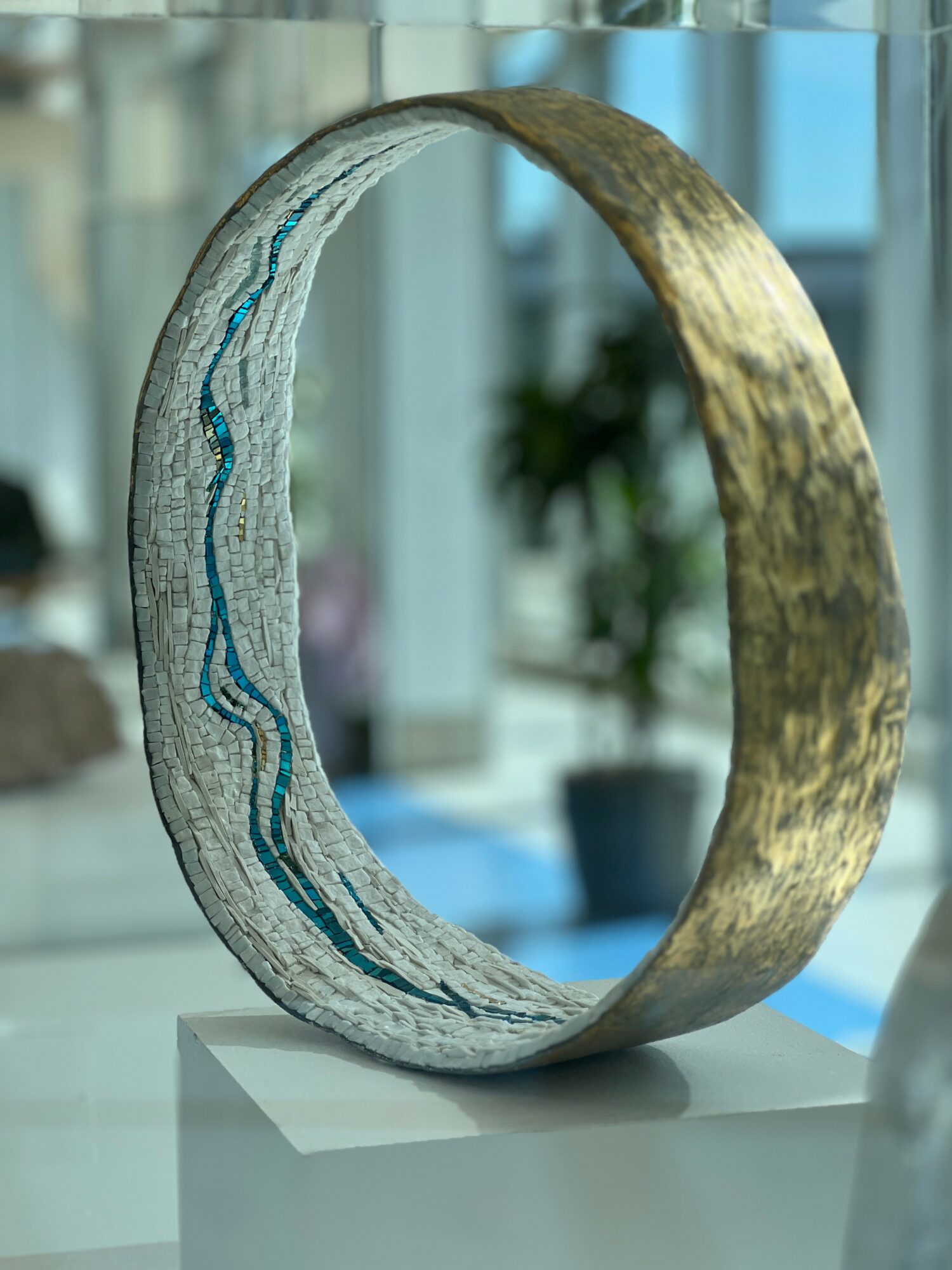 A circular metallic sculpture with textured surface and blue lines, displayed indoors with blurred background of plants and windows.