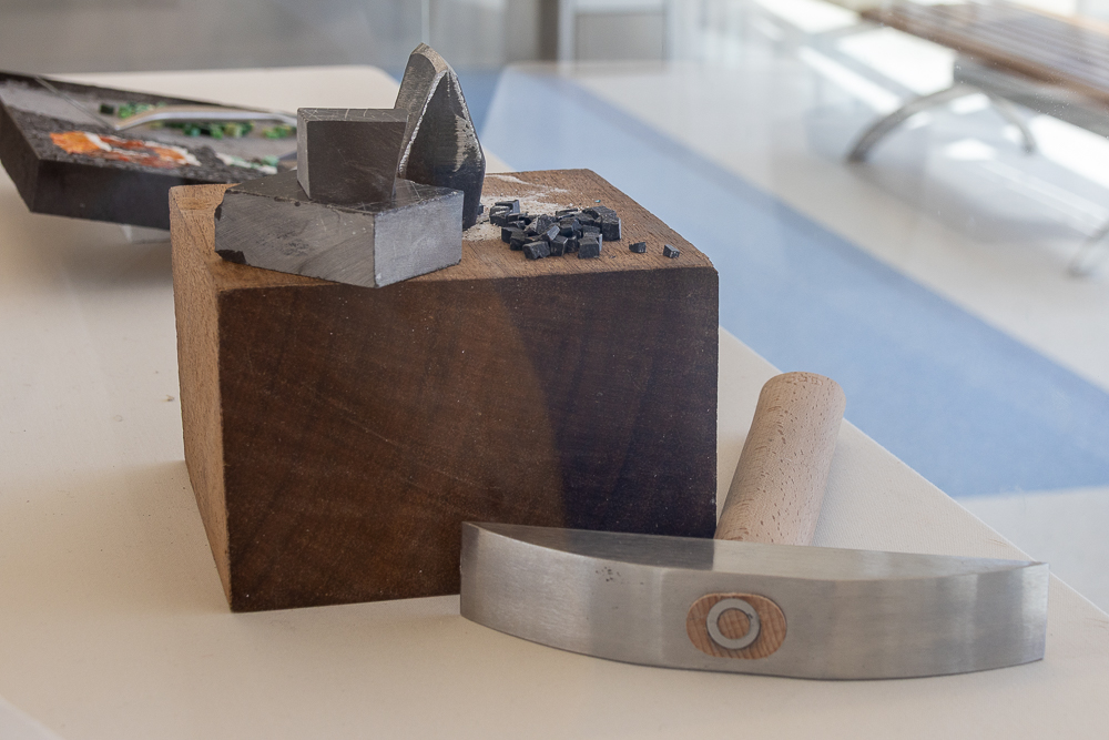 Wooden block with gray and black objects on top, a curved metal tool in front, and a rolling pin nearby, on a white surface.