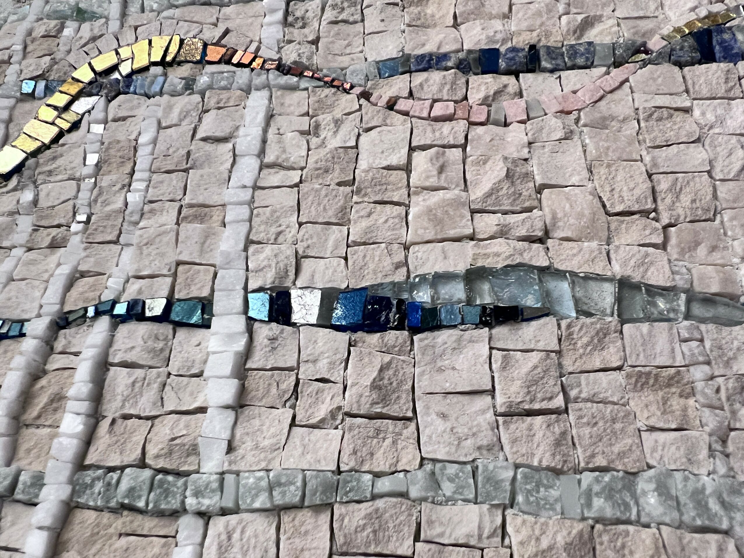Colorful mosaic snake on cobblestone ground, with various shades of yellow, blue, and gray tiles, curving across the surface.