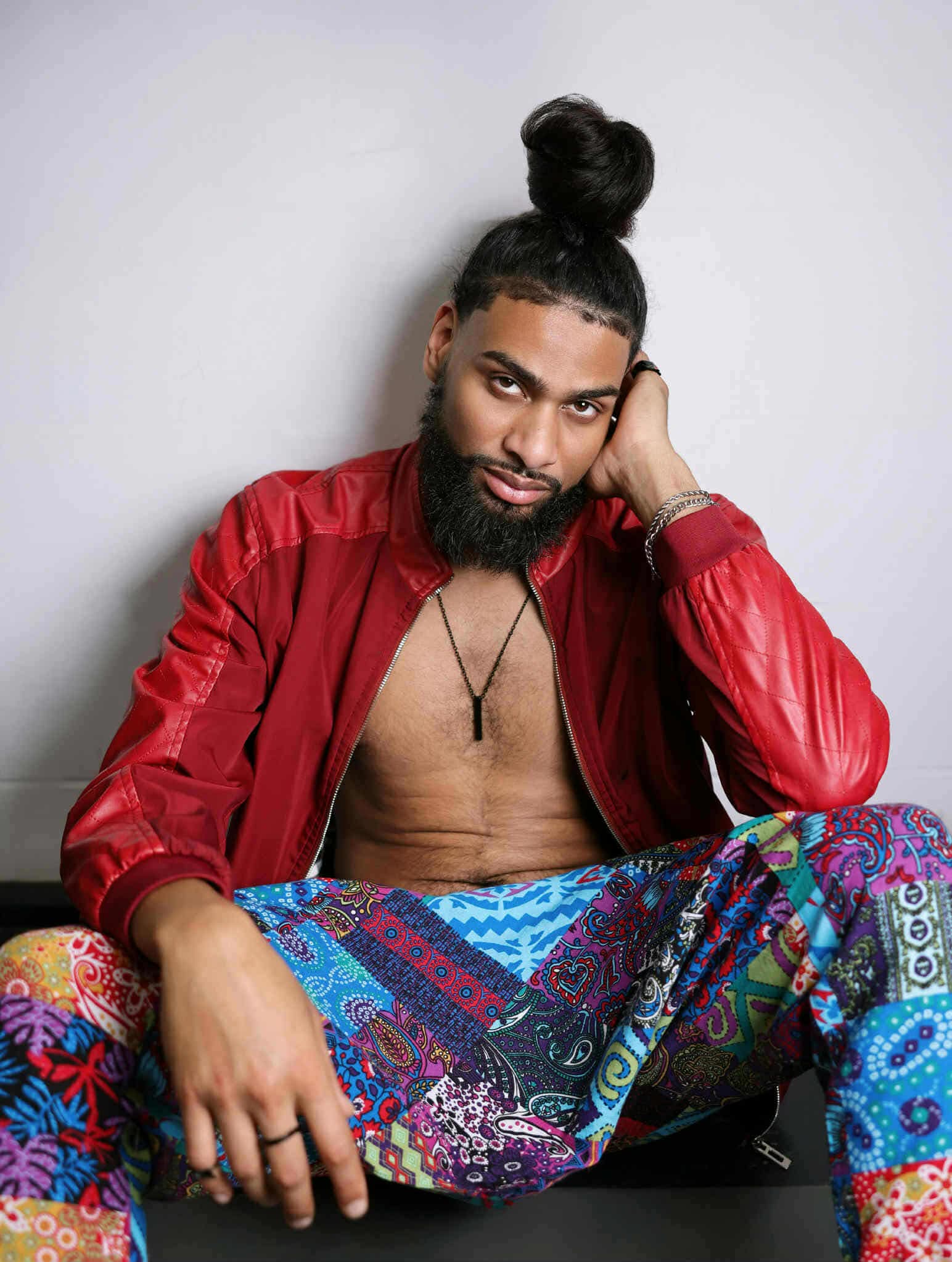 Man with a top knot hairstyle, beard, red jacket, colorful patterned pants, sitting against a plain background.