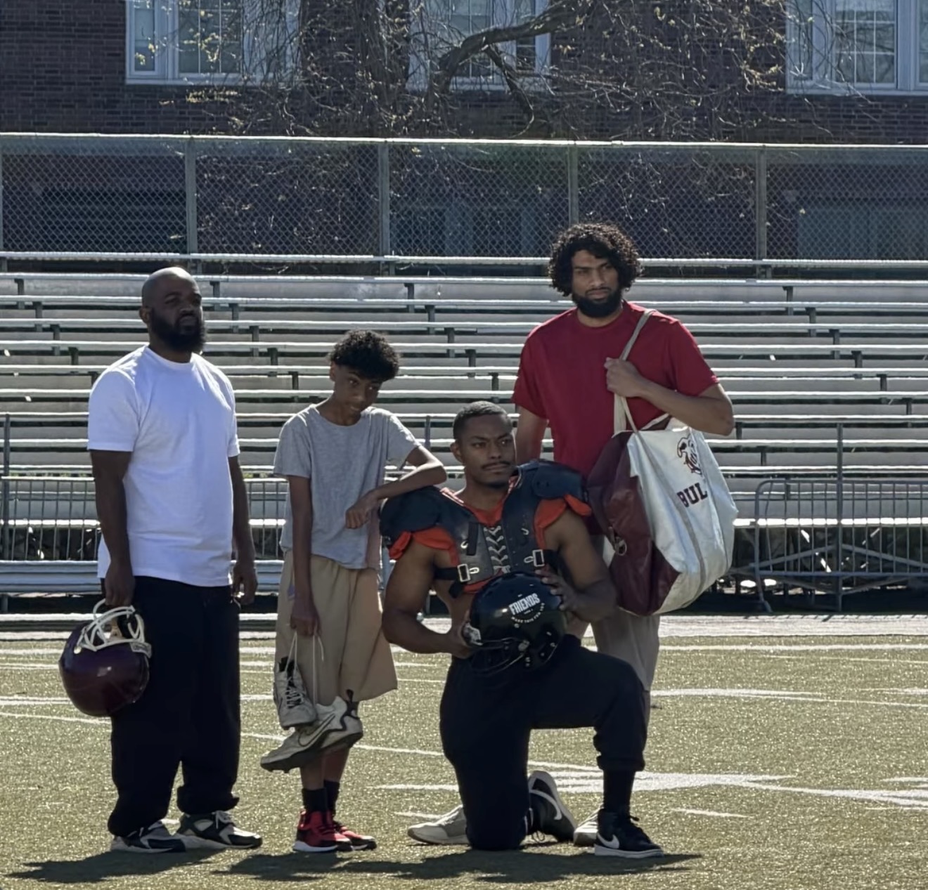 Four people standing on a sports field with bleachers in the background, one holding a helmet, one with a football, and two carrying bags.