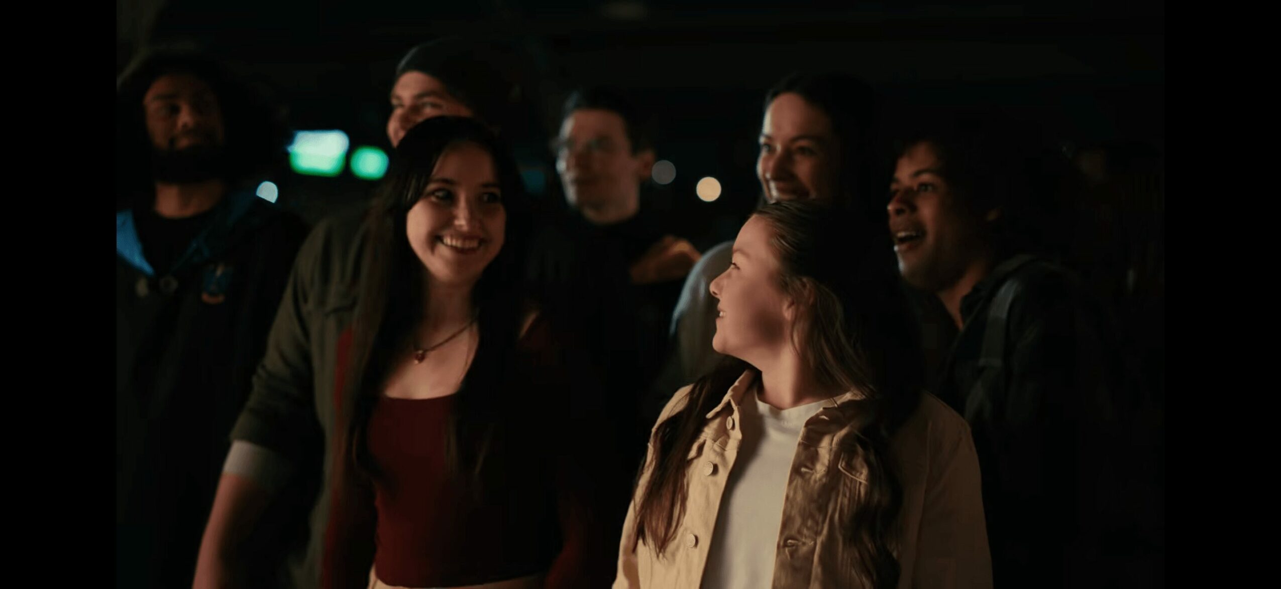 Group of people smiling and talking in a dark setting, some illuminated by soft light, with a focus on two women in the foreground.