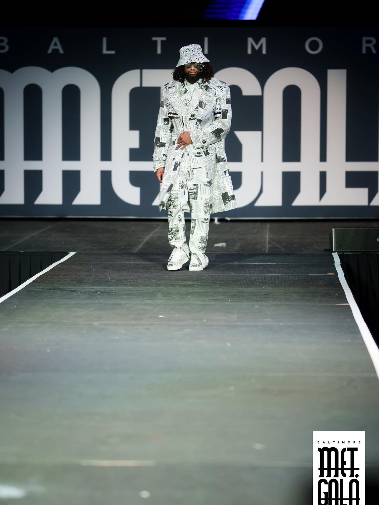 Person walking on runway wearing a patterned outfit and hat, with large text background, at a fashion event.