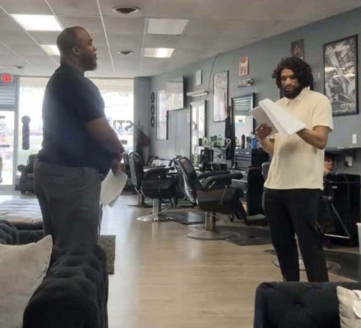 Two men stand and talk in an office, one holding a paper, the other reading a book, with desks and chairs behind them.