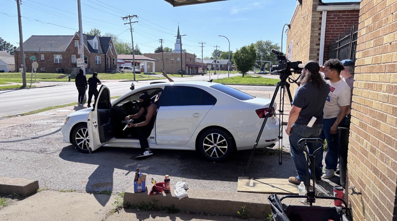 People filming a white car parked on the street, with equipment and supplies on the ground, near a brick building.