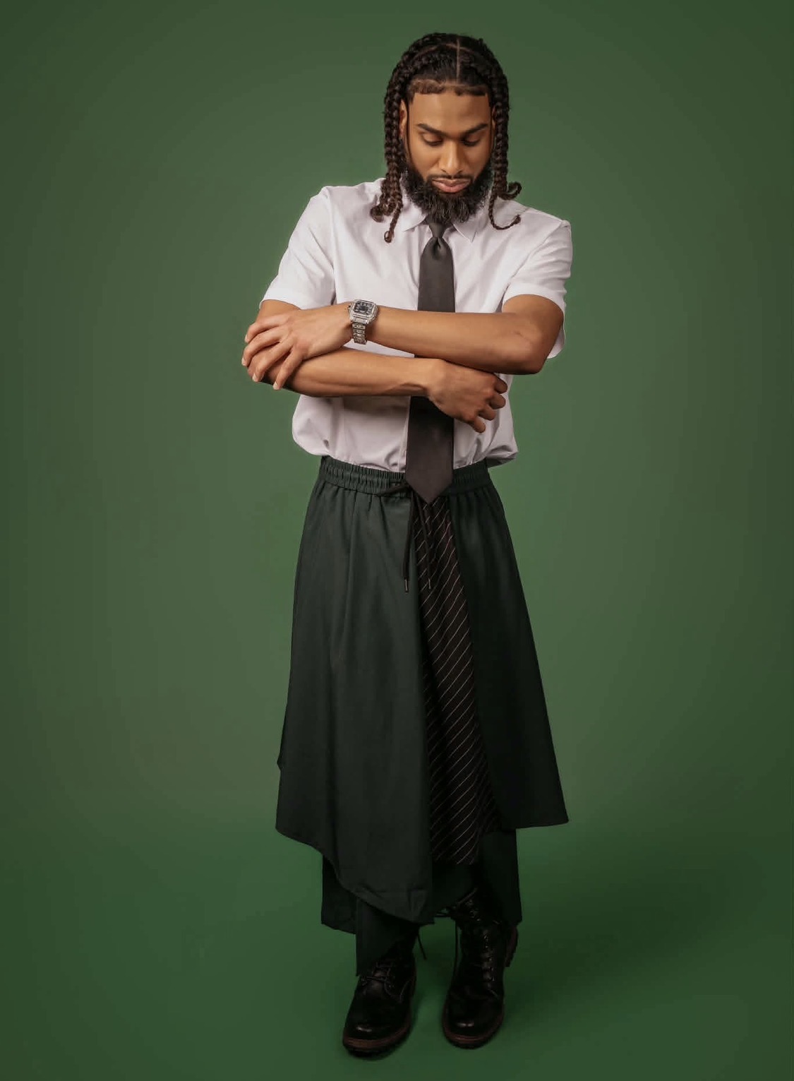Man with dreadlocks wearing a white shirt, black tie, and long black skirt, standing against a green background.