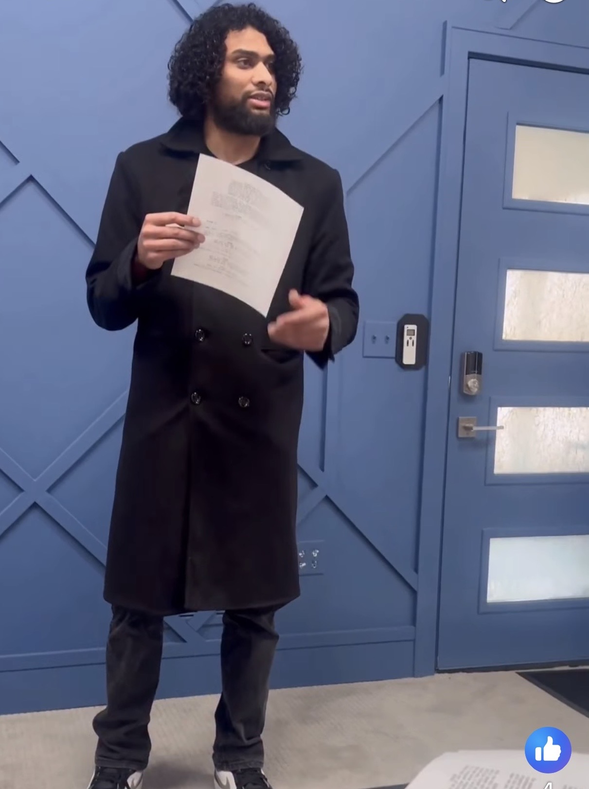 Man with curly hair and beard holding a paper, standing in front of a blue wall and door.