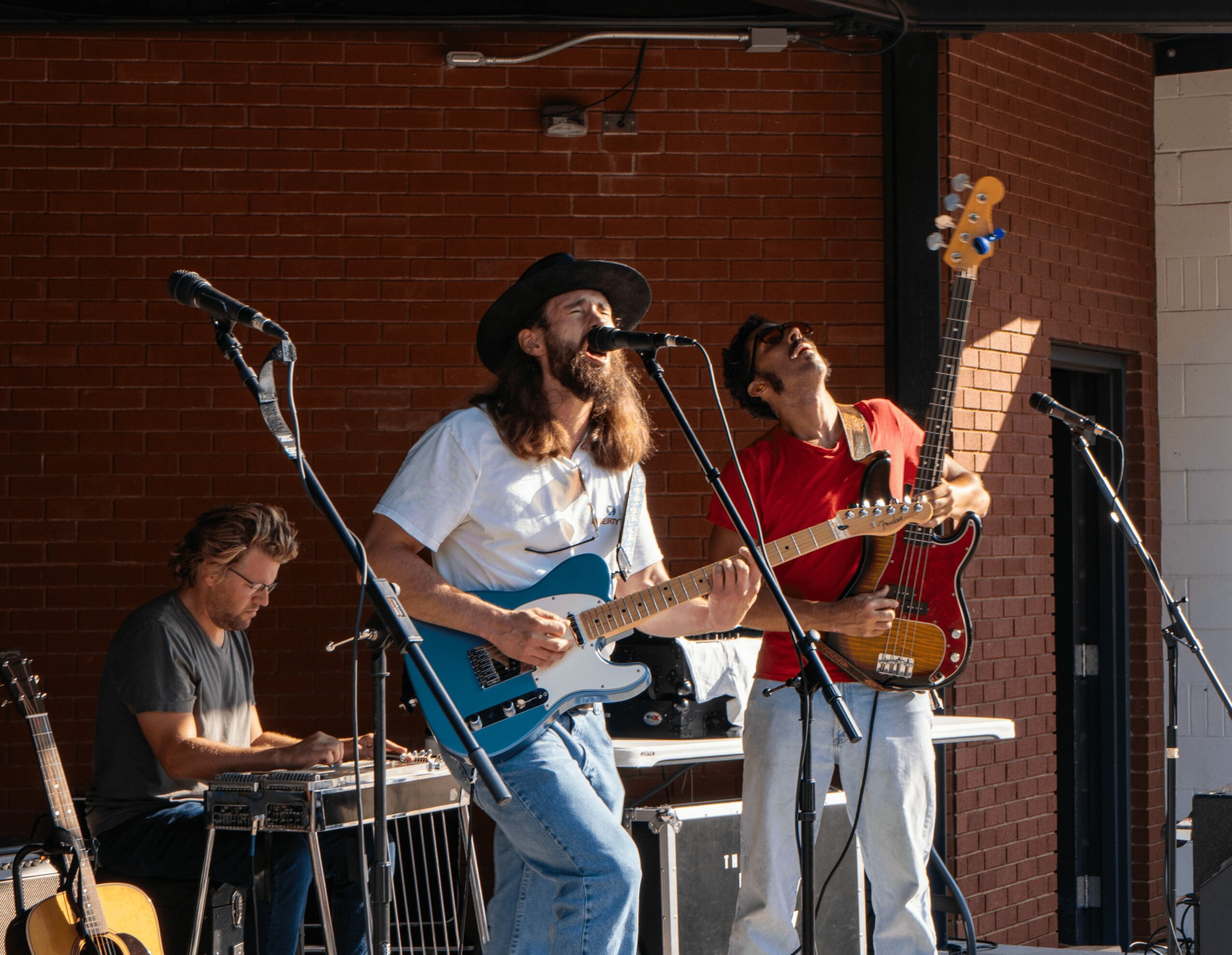Three musicians perform outdoors against a brick wall, playing guitar, keyboard, and bass guitar.