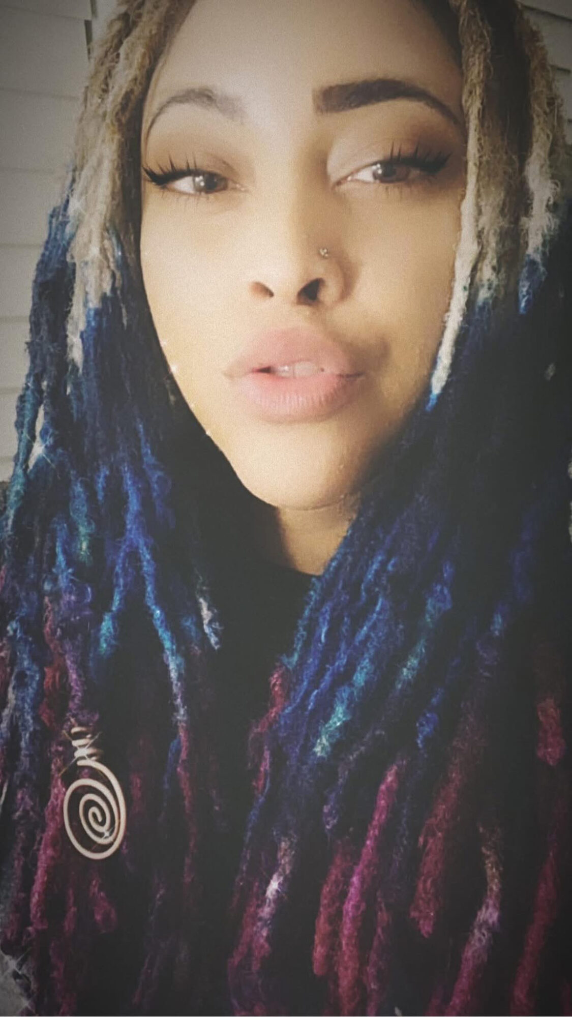 Close-up of a woman with multicolored dreadlocks and makeup, looking at the camera with a neutral expression.