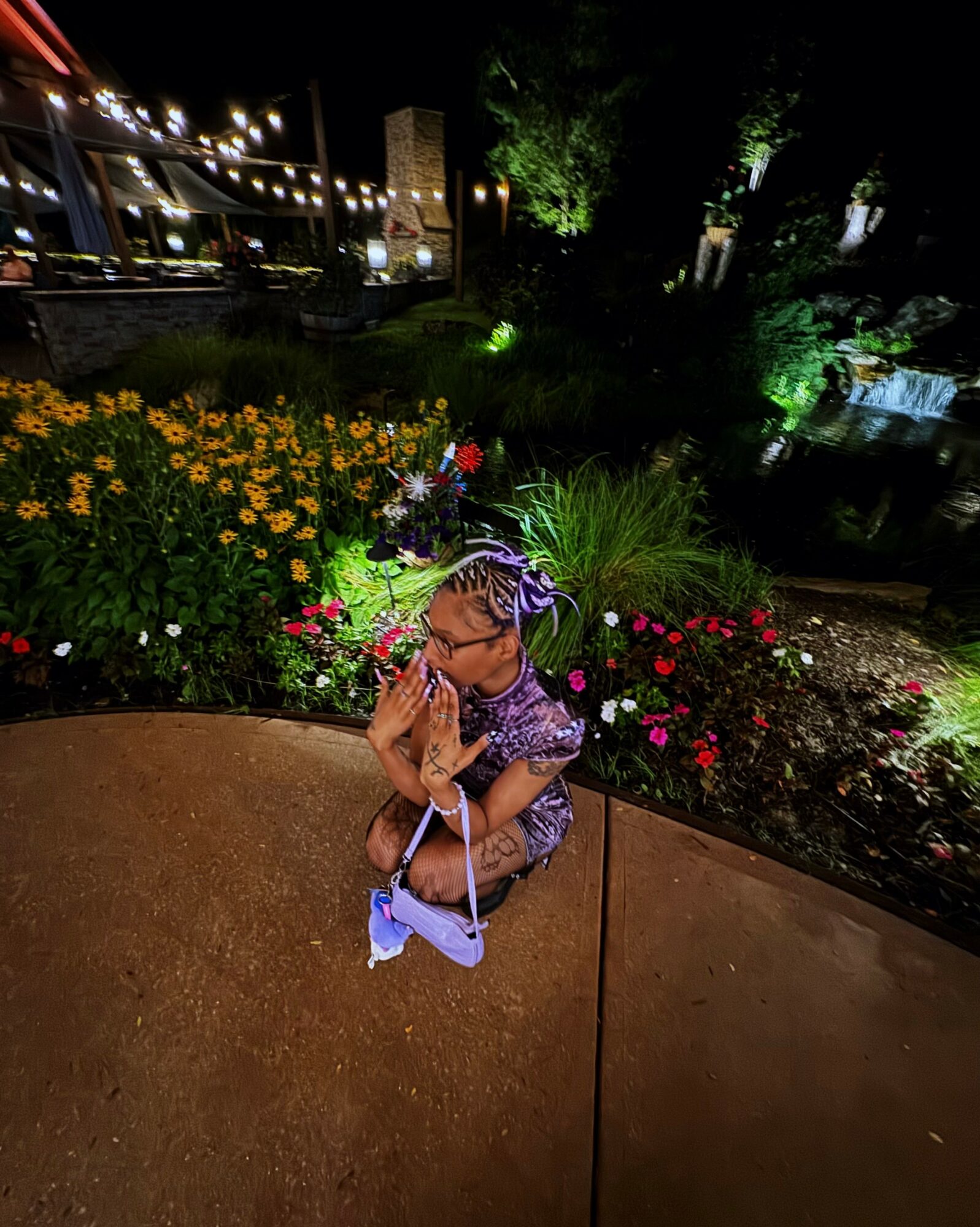 Child wearing a colorful hat and glasses, sitting outdoors near flowers and greenery at night, illuminated by lights.
