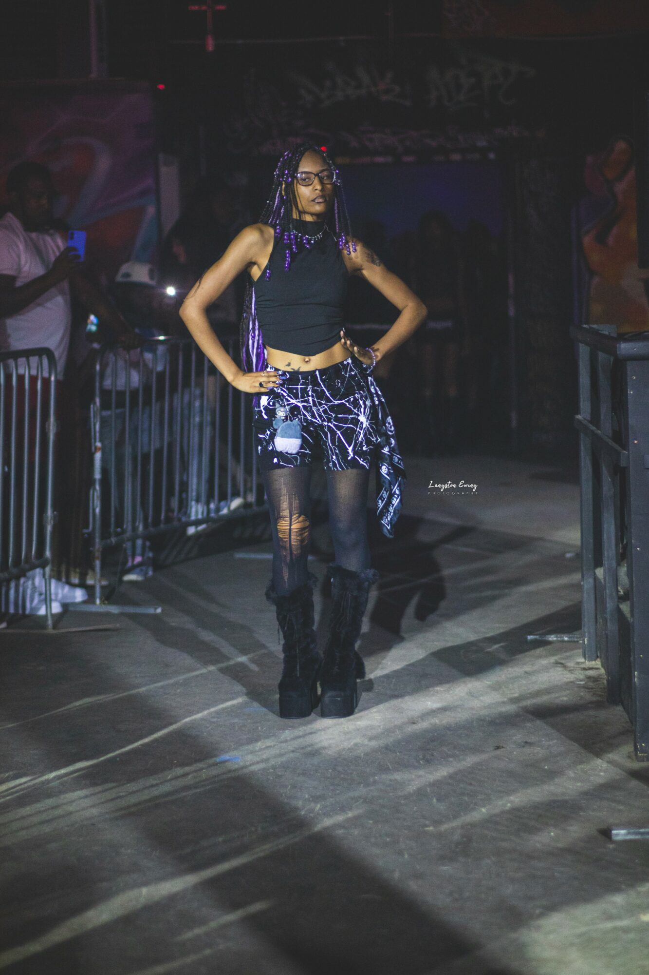 Person walking on a runway with hands on hips, wearing dark clothing and accessories, in a dimly lit environment.