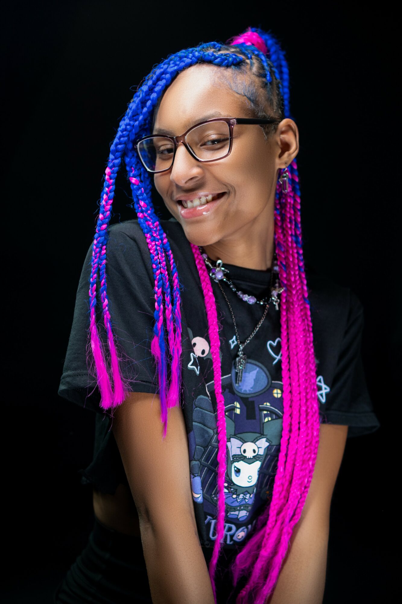 Young person with colorful braided hair, glasses, smiling, wearing a black graphic t-shirt, against a dark background.