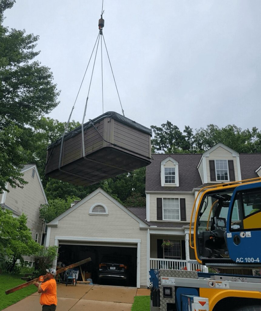 A crane lifts a large storage shed above a residential house with a garage, trees, and cloudy sky in the background.