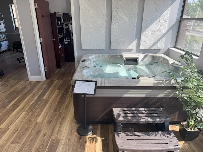 Indoor hot tub with control panel, next to a potted plant, in a room with large windows and wooden flooring.