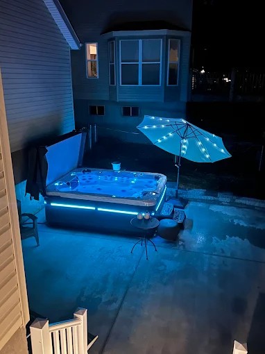 Hot tub with blue LED lights, an illuminated umbrella, and a house in the background at night.