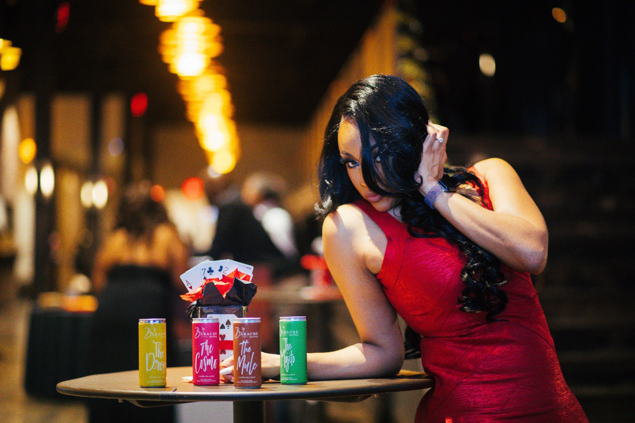 Woman in red dress leaning on a table with colorful cans, in a dimly lit setting with blurred background