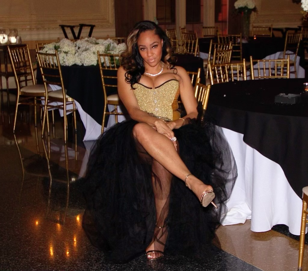 Young woman in a gold and black dress sitting on a chair at a decorated event with tables and chairs in the background.