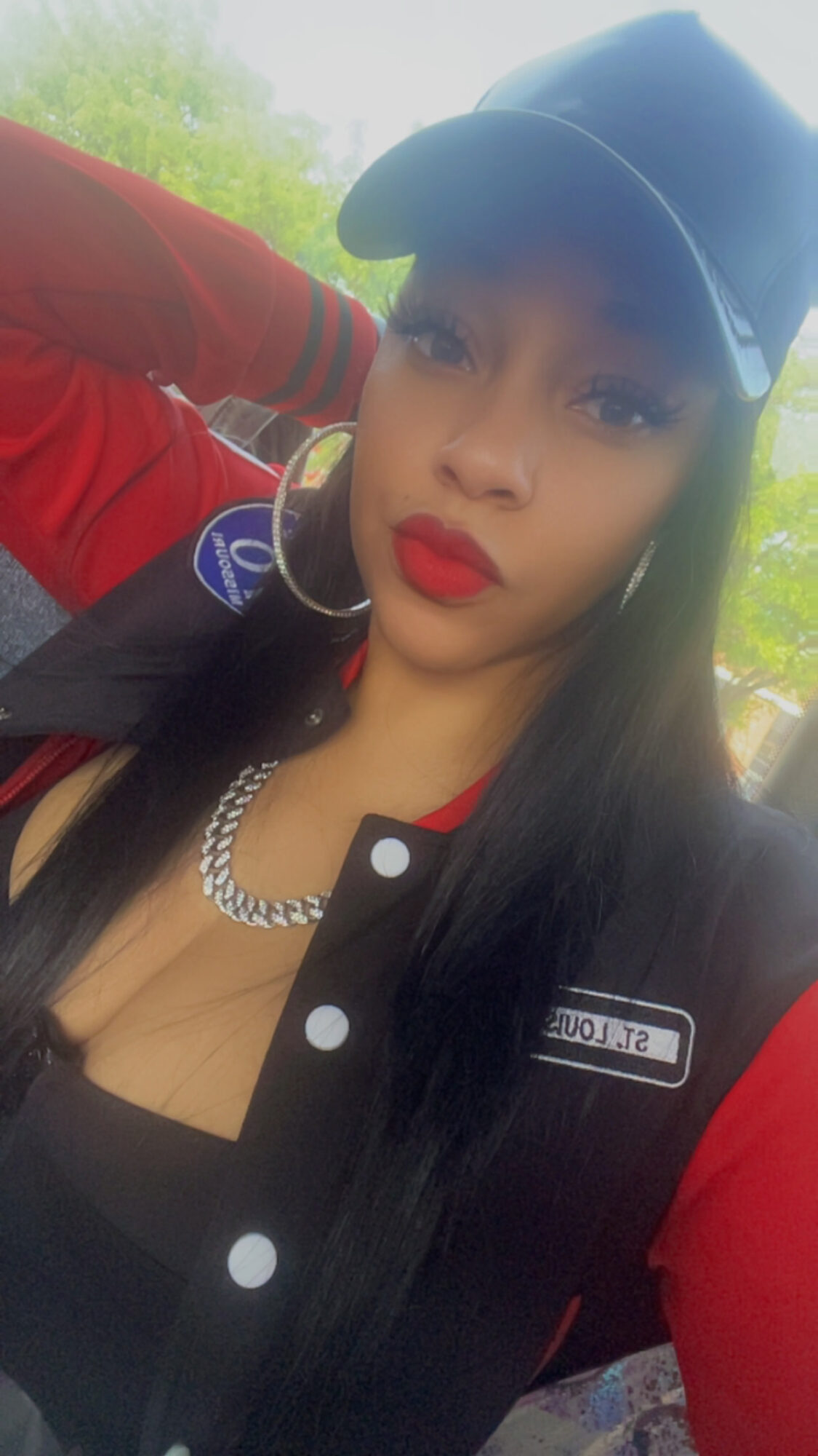 Young woman with long black hair wearing a blue cap, red lipstick, and a black and red jacket outdoors.