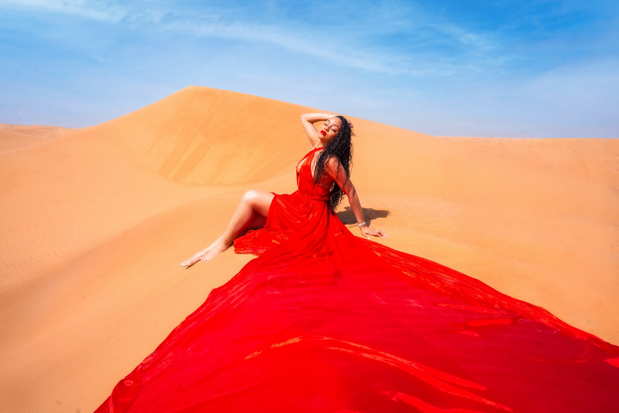 Woman in a red dress sitting on sand dunes under a blue sky, with her head tilted back and hand on her forehead.
