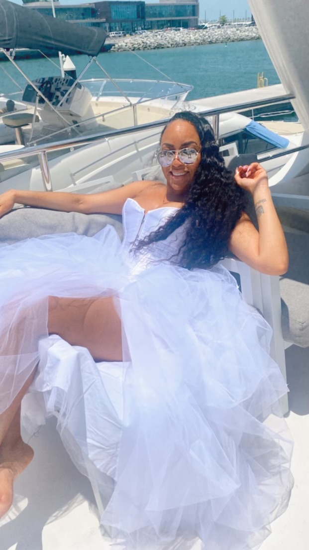 Woman in a white dress relaxing on a boat with water and other boats in background.