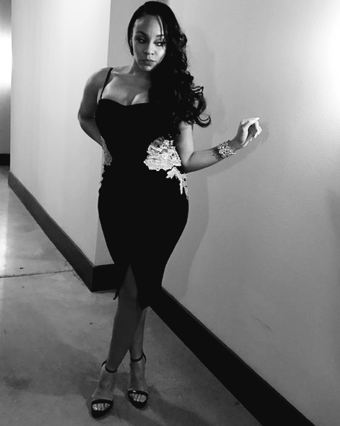 Woman in a black dress with floral details, standing in a hallway, looking at the camera, with long curly hair.
