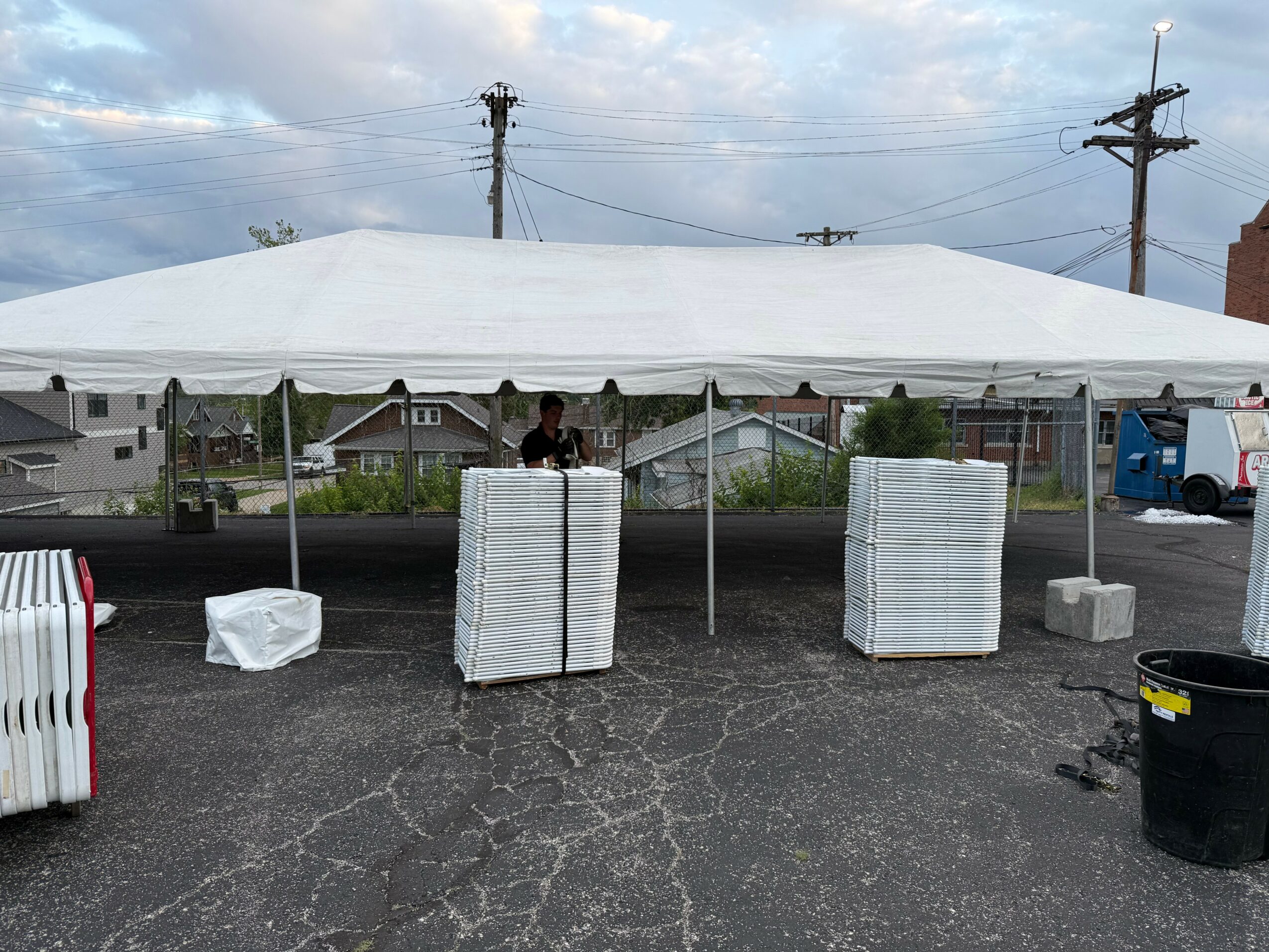 White canopy tent set up outdoors with stacks of white pallets underneath, on a cracked asphalt surface, with power lines and buildings in background.