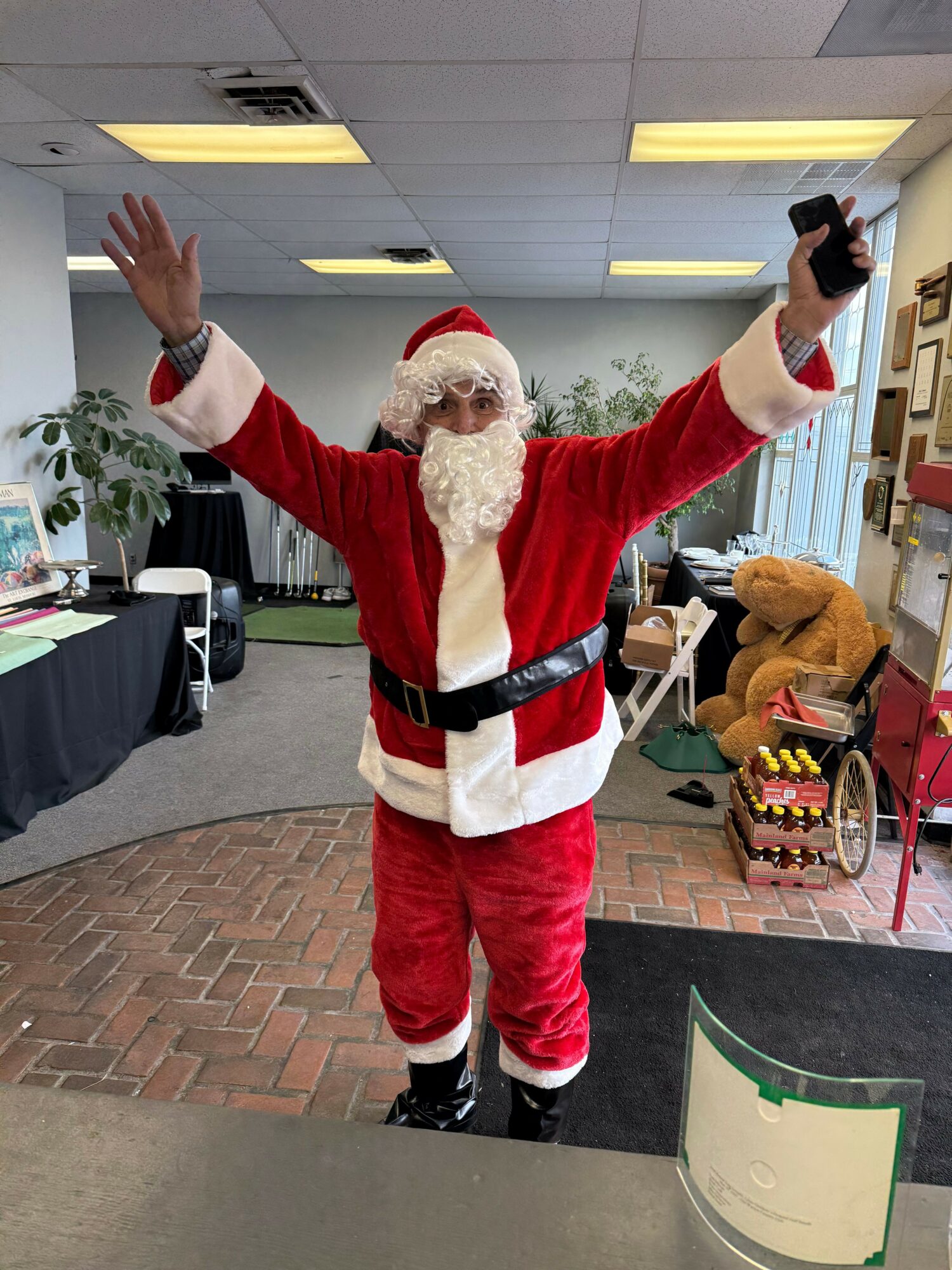 Santa Claus in a red suit with white trim, waving and holding a phone, standing indoors with a brick floor.