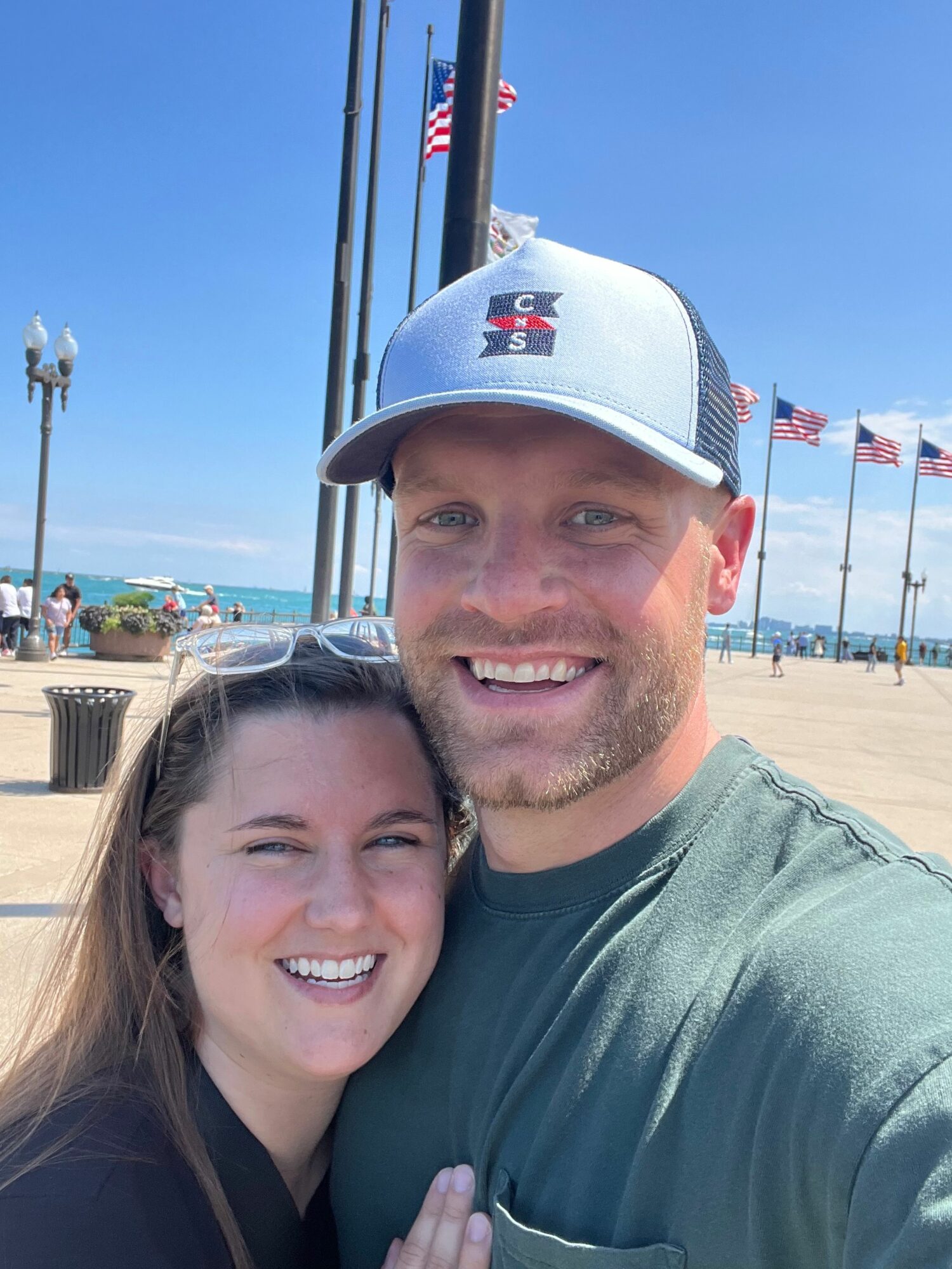 Two people smiling at the camera, outdoors with a beach and flags in the background.