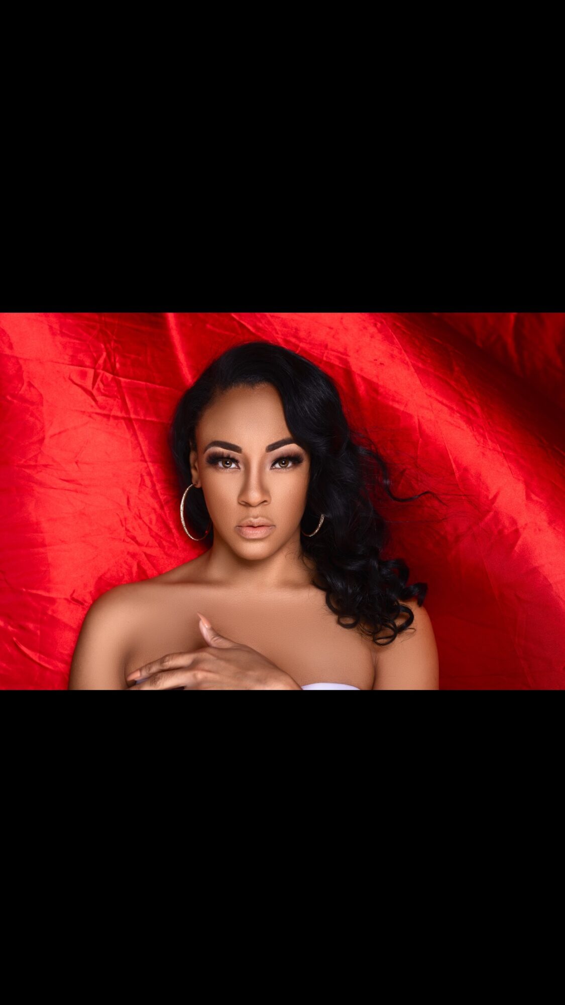 Portrait of a woman with dark hair and hoop earrings, posing against a red fabric background.