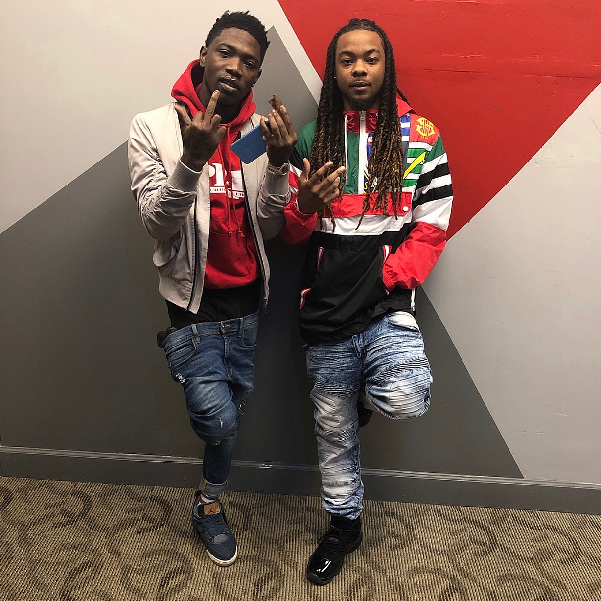 Two men standing against a wall with geometric red, gray, and white design, both making hand gestures, wearing casual clothing.
