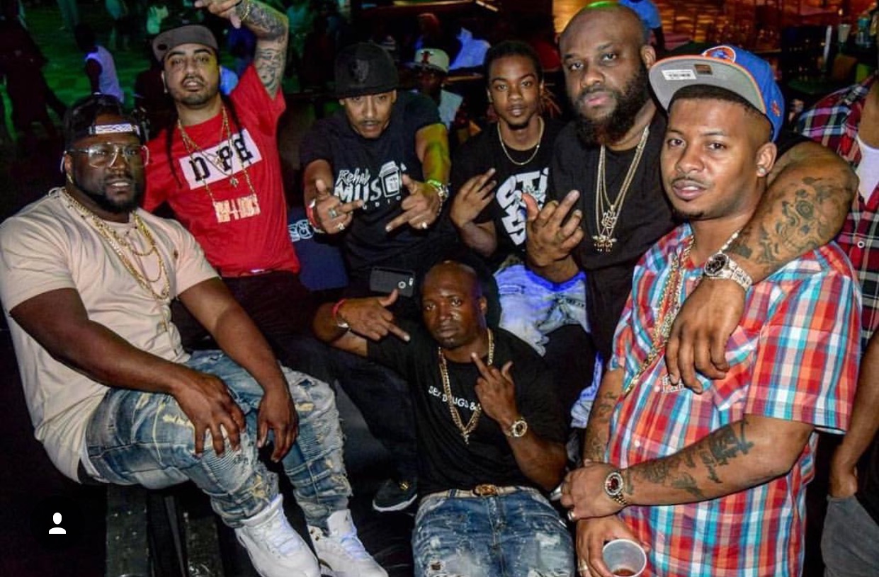 Group of seven men posing together in a lively indoor setting, some making hand gestures, wearing casual clothing and accessories.