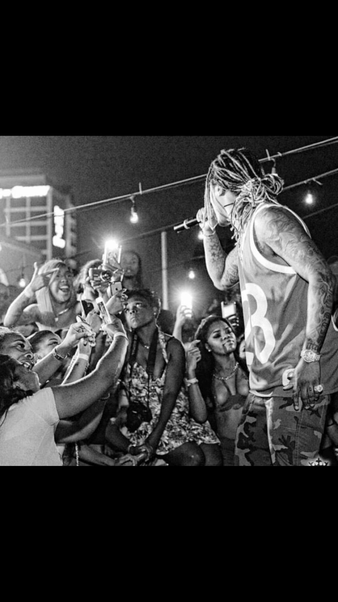 A performer with braided hair and tattoos interacts with a group of children at night under string lights.