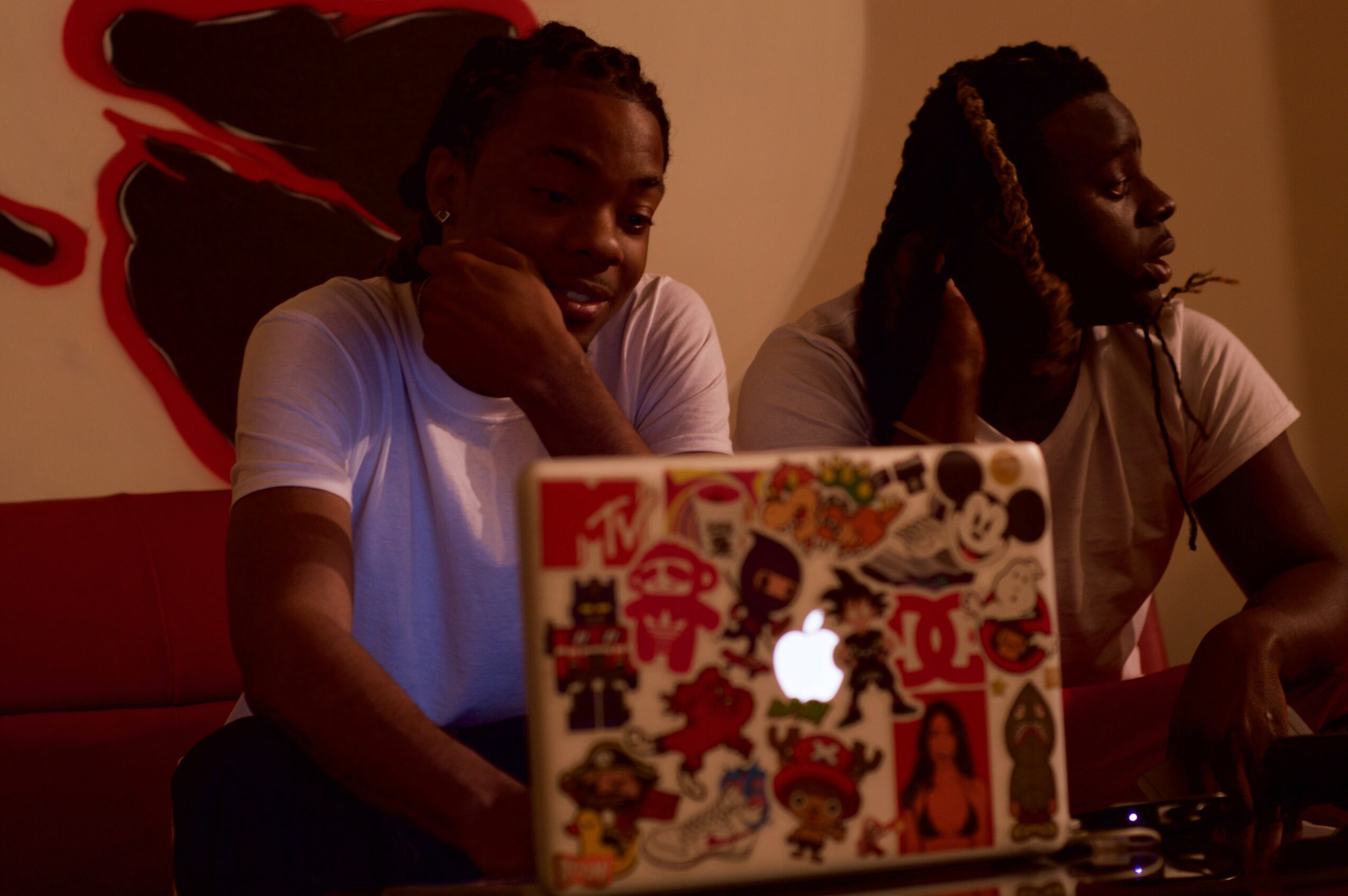 Two people sitting at a table with a decorated laptop, one resting their head on their hand, the other looking away.