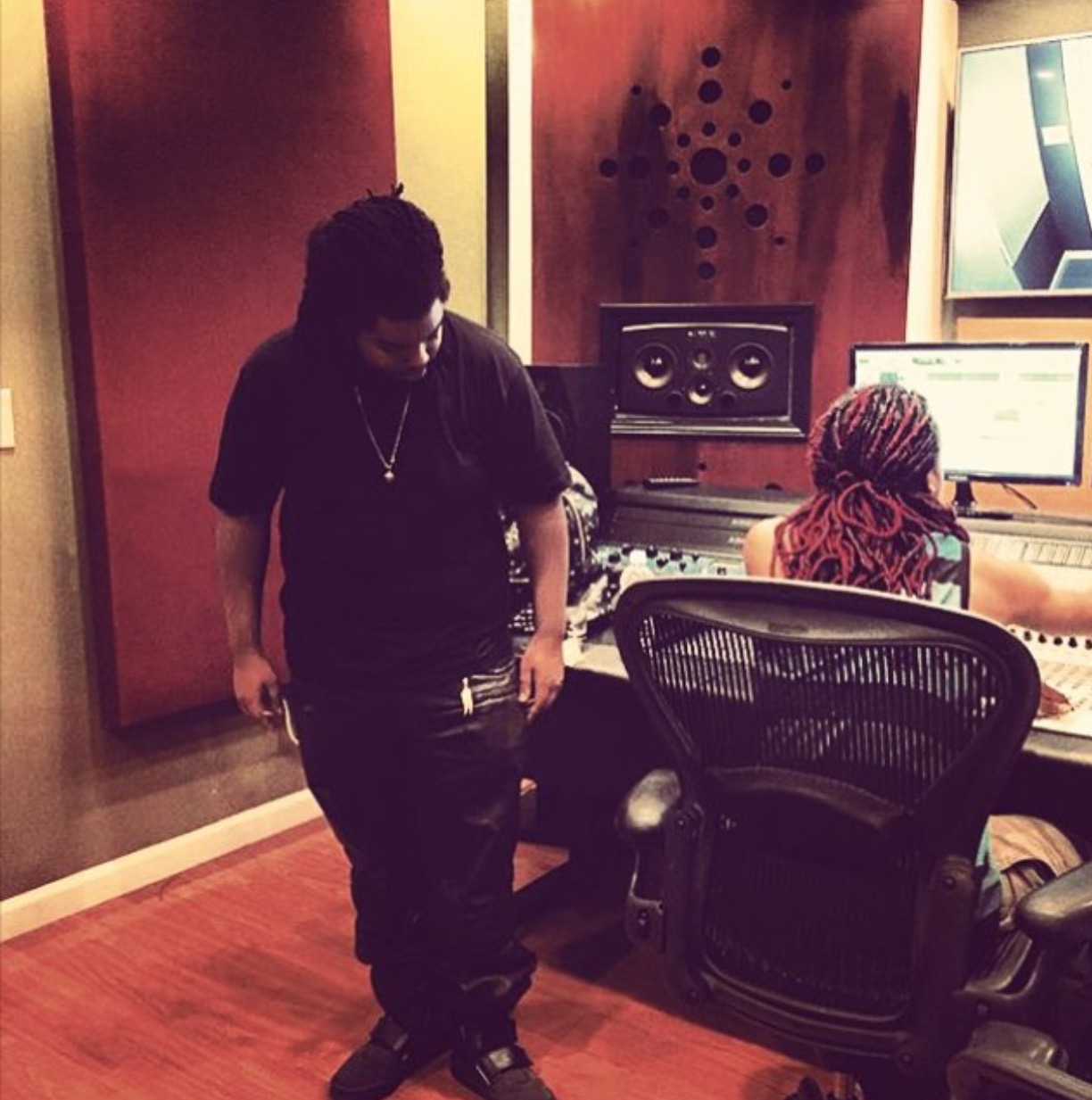 Two people in a recording studio, one standing and one sitting at a mixing console, with audio equipment and a monitor.