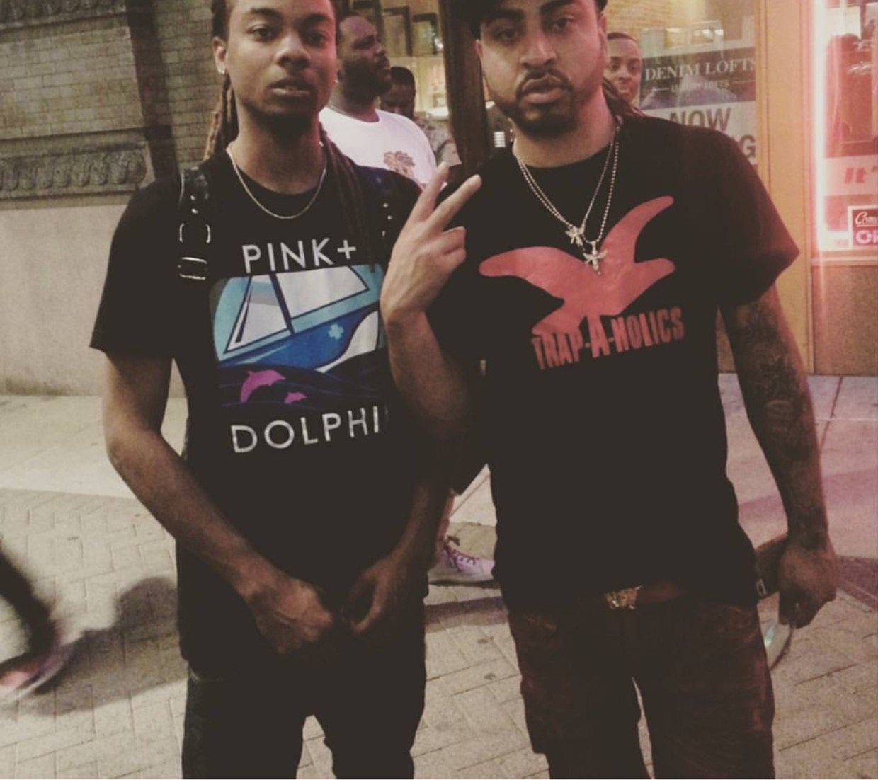 Two young men standing outdoors, one making a peace sign, both wearing black T-shirts with graphic designs.