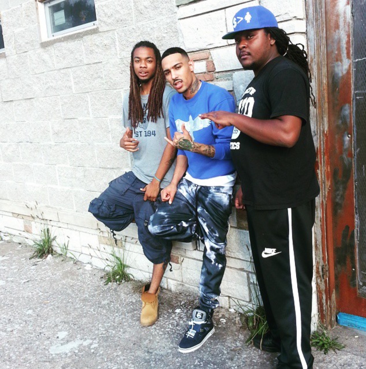 Three people standing outdoors against a brick wall, one reading a book, two looking at the camera.
