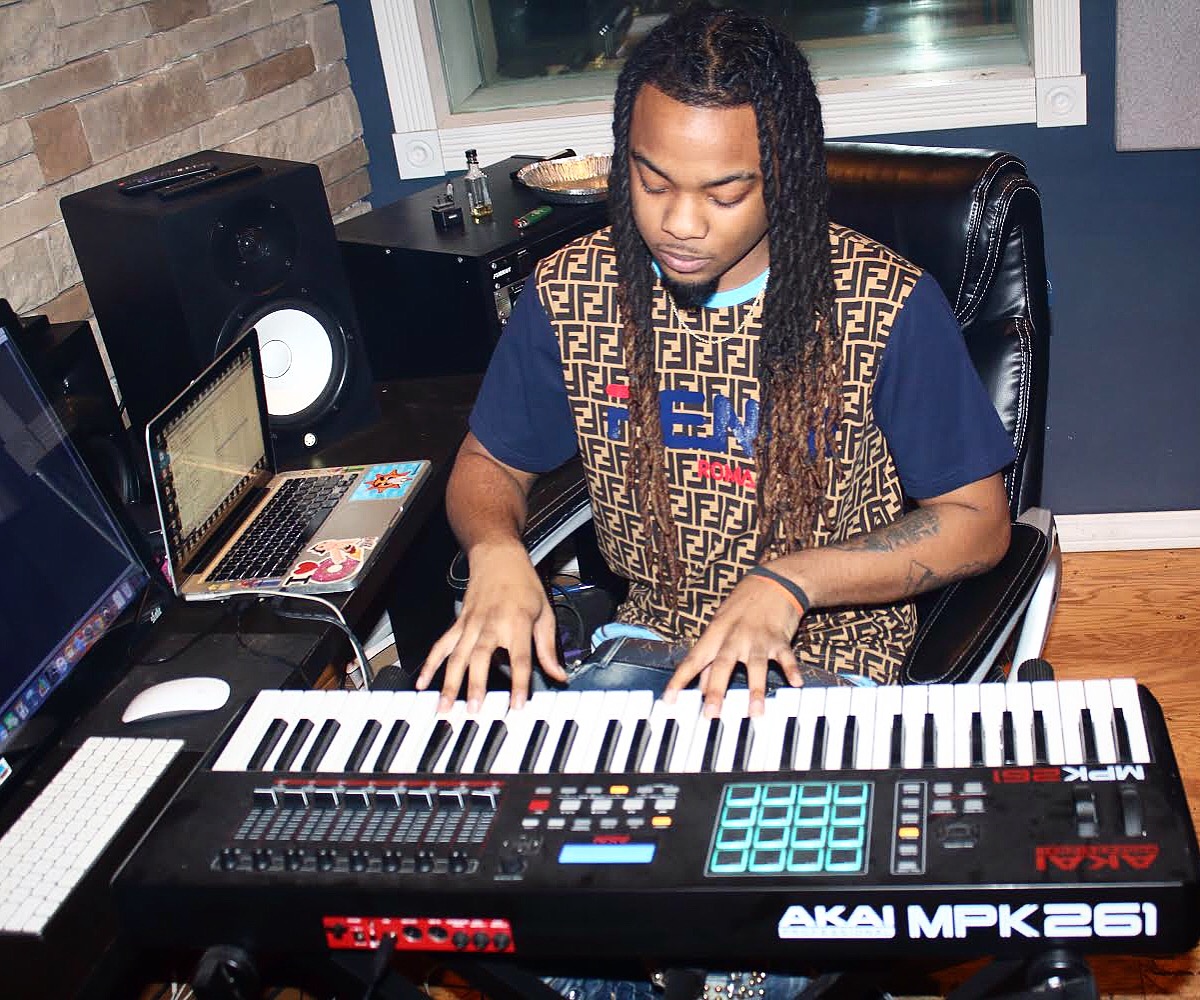 Person with long braided hair playing a keyboard in a music studio, surrounded by audio equipment.