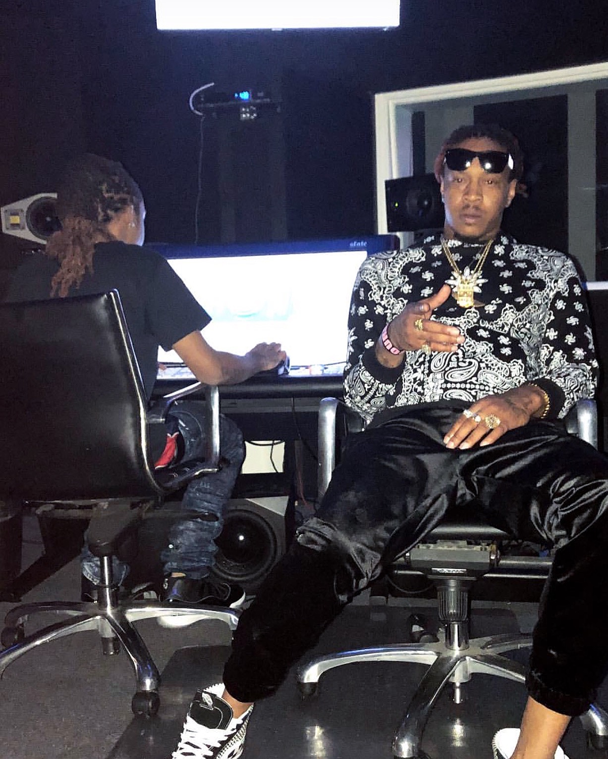 Person wearing sunglasses and patterned shirt sitting in a recording studio, holding a phone, with another person working on a computer in the background.