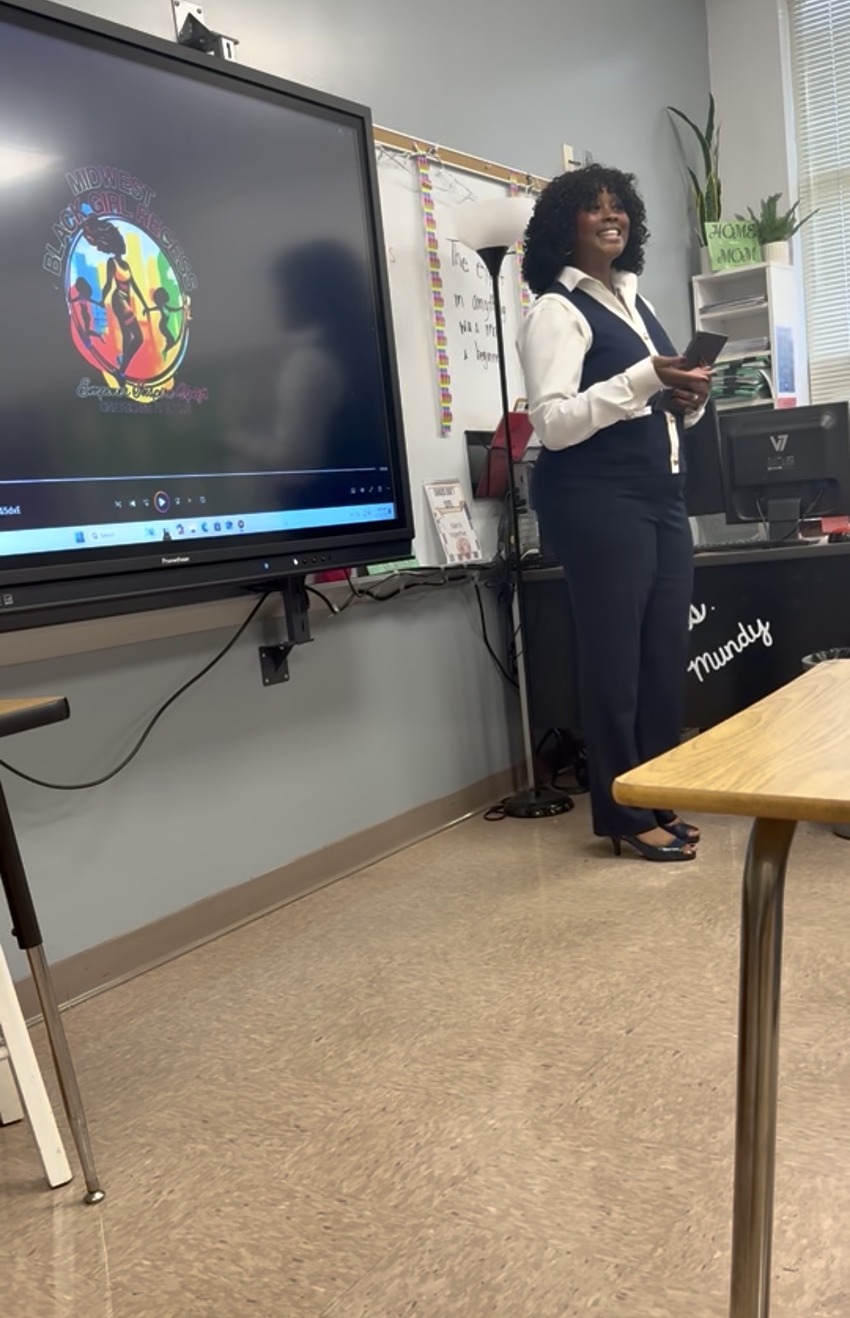 Woman with curly hair and glasses stands in front of a classroom, holding a tablet, with a large screen displaying a colorful logo behind her.