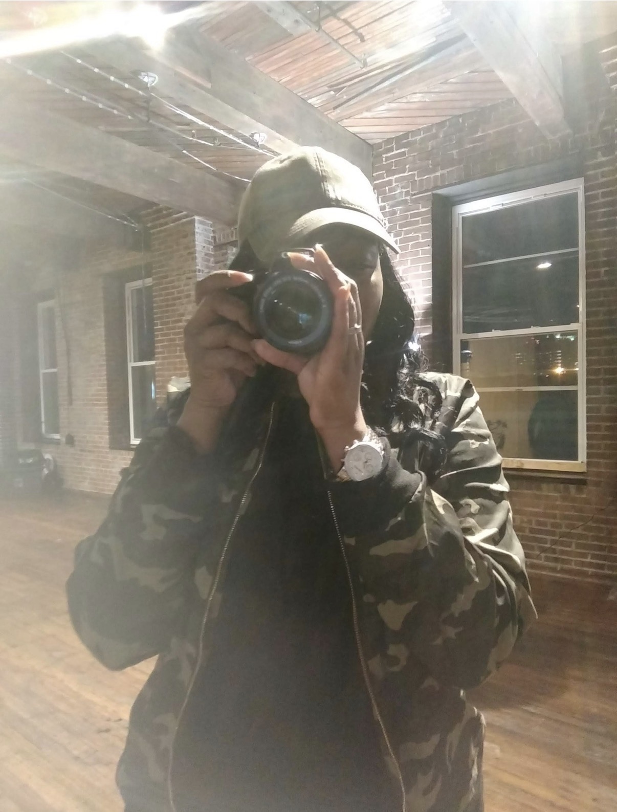 Person taking a photo in a room with brick walls and windows, wearing a cap and camouflage jacket.