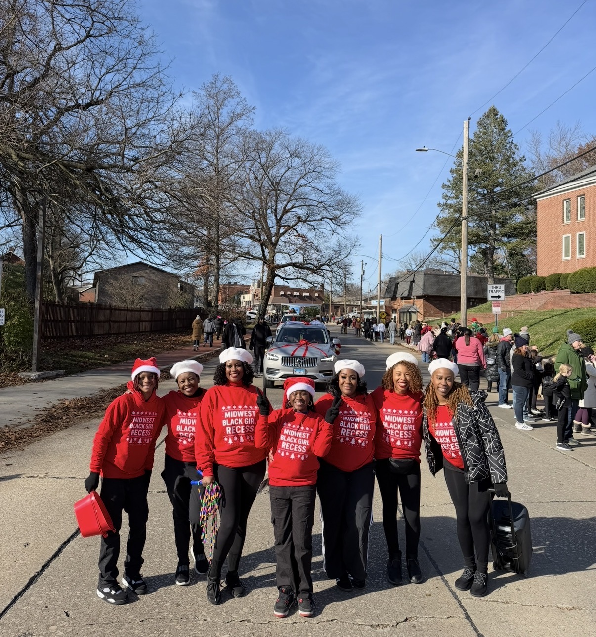 Group of children wearing red shirts and Santa hats walking outdoors on a sunny day, some holding bags or containers.