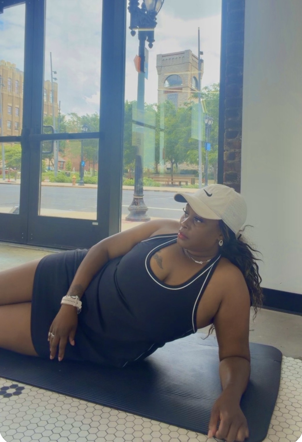 Woman lying on a mat indoors, wearing a black tank top and white cap, looking outside through large windows.