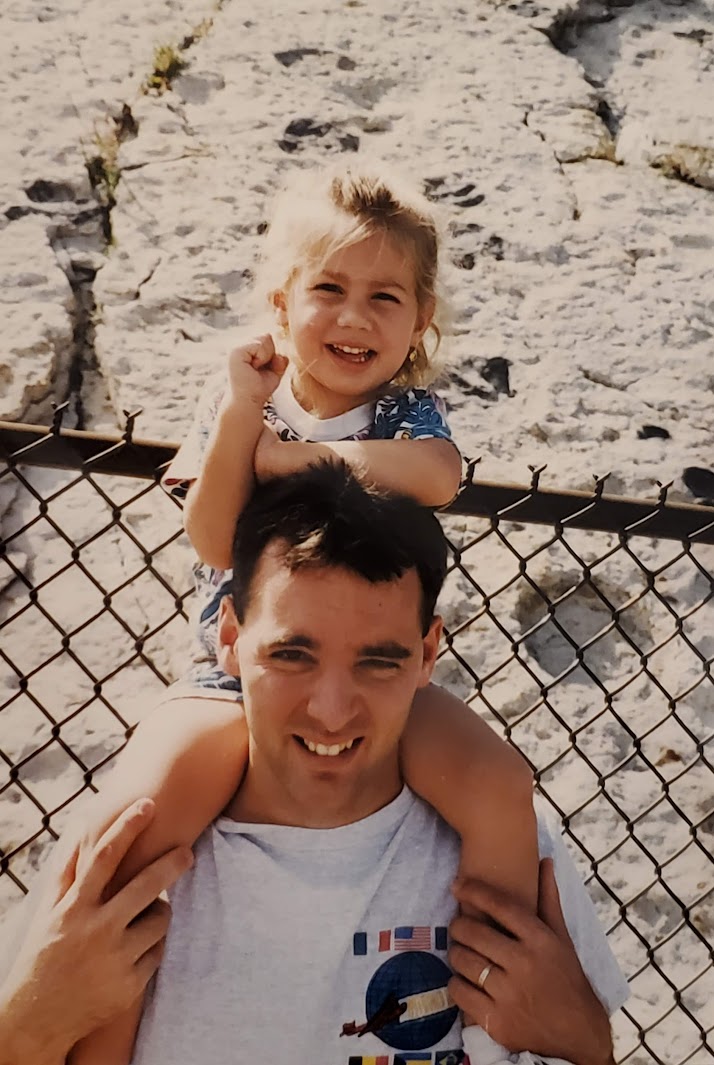 Child sitting on man's shoulders smiling, outdoors with rocky background and fence.