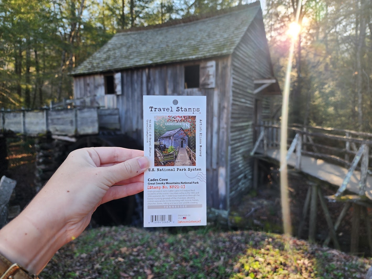 Hand holding a travel stamp with a rustic wooden building and trees in the background, sunlight shining through.