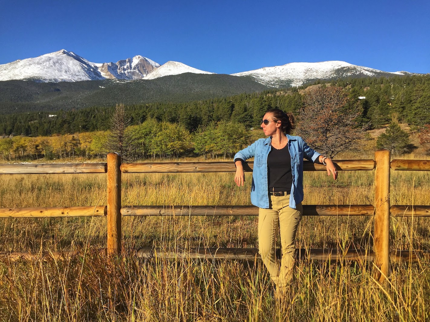 Woman standing by a wooden fence with mountains and trees in the background, wearing sunglasses and casual clothing.