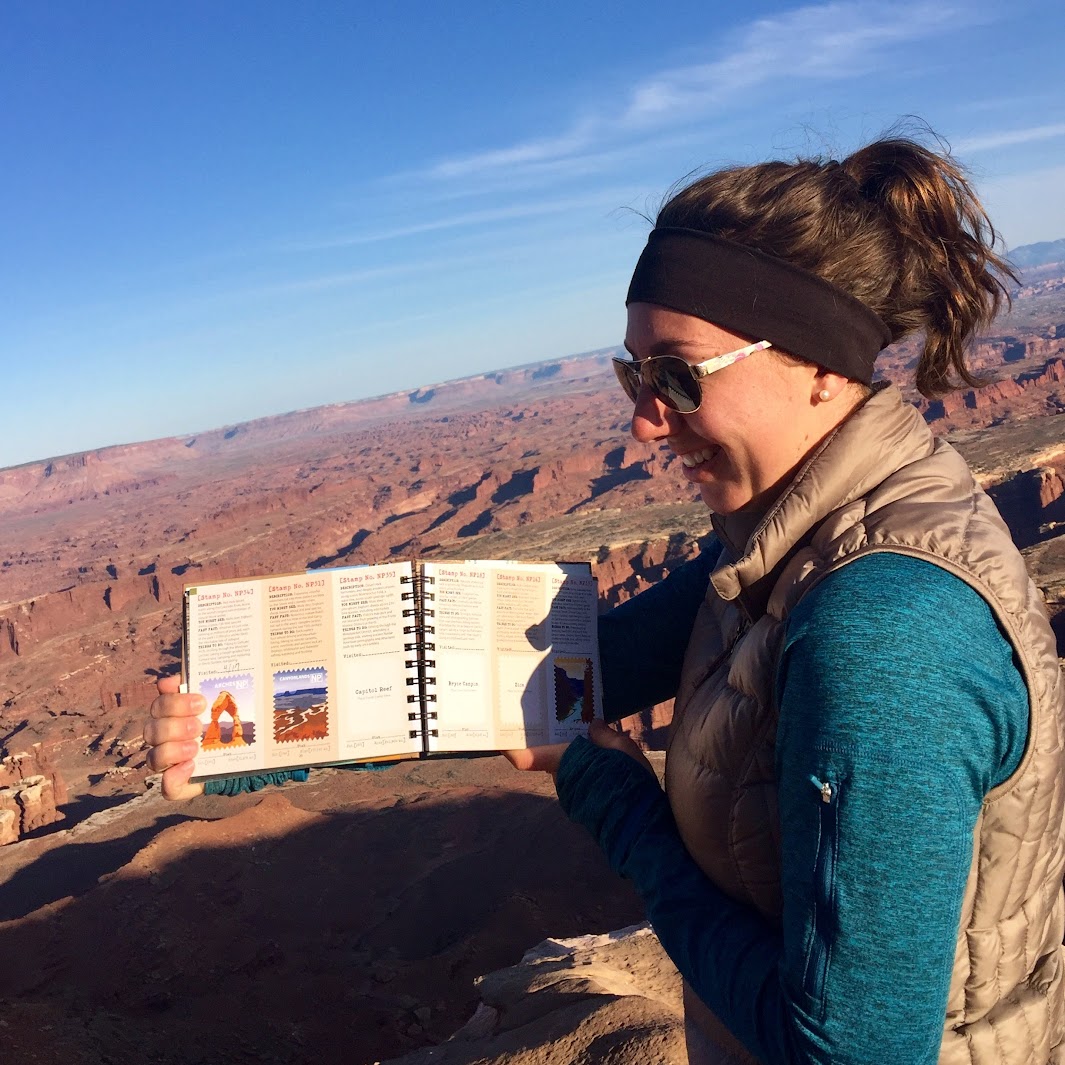 Woman with sunglasses, headband, and vest reading a guidebook outdoors with canyon landscape in background.