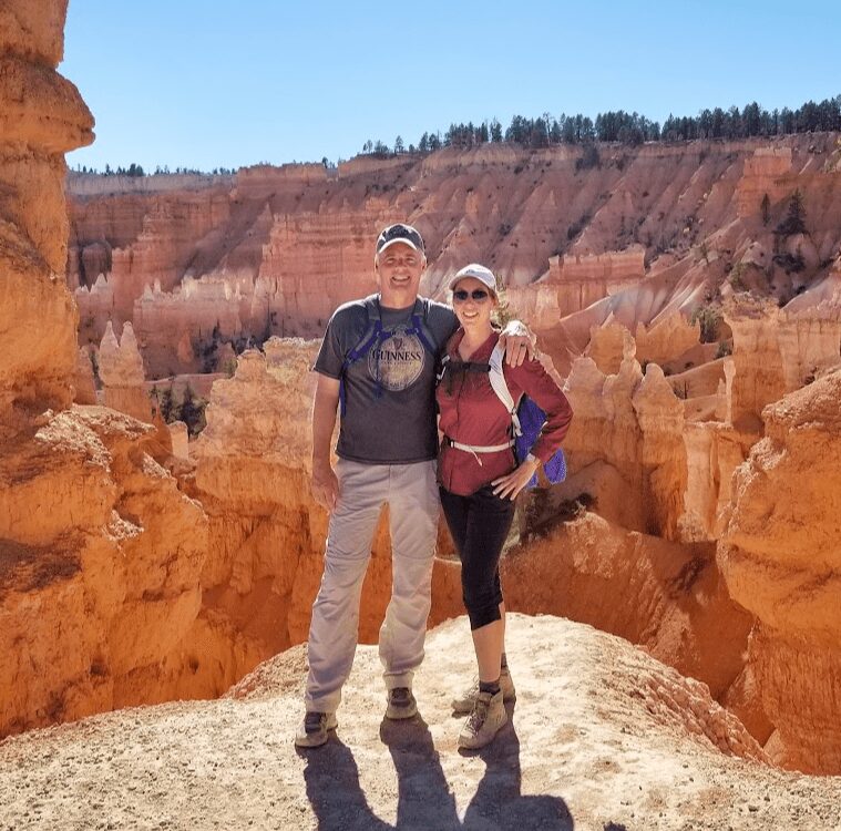 Two people stand on a rocky ledge with colorful canyon formations in the background, smiling at the camera.