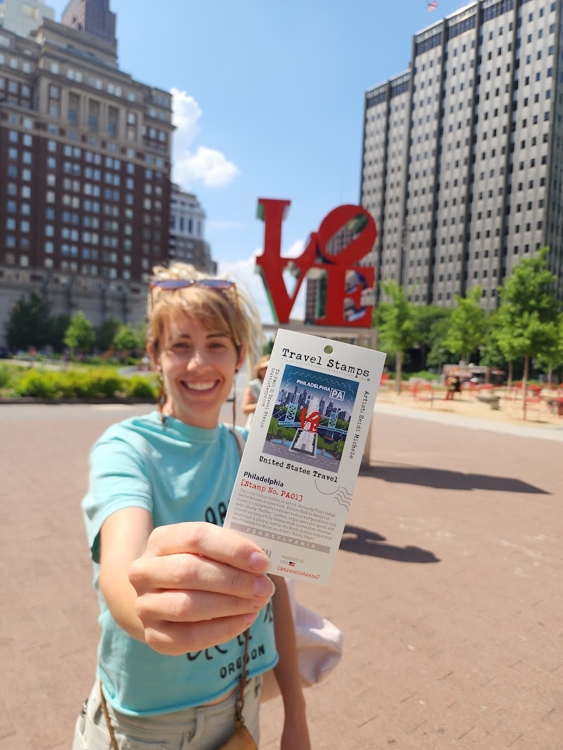 Smiling young woman holding a travel stamp in an outdoor urban park with tall buildings and a red 'LOVE' sculpture behind her.
