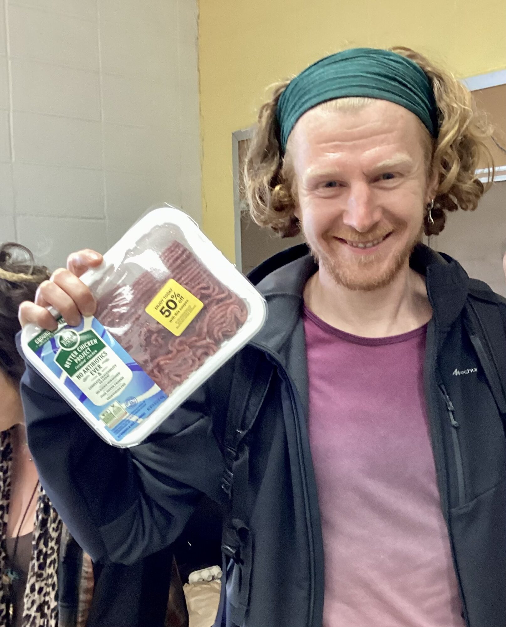 Person with curly hair, headband, smiling, holding packaged ground meat, indoors with yellow wall background.